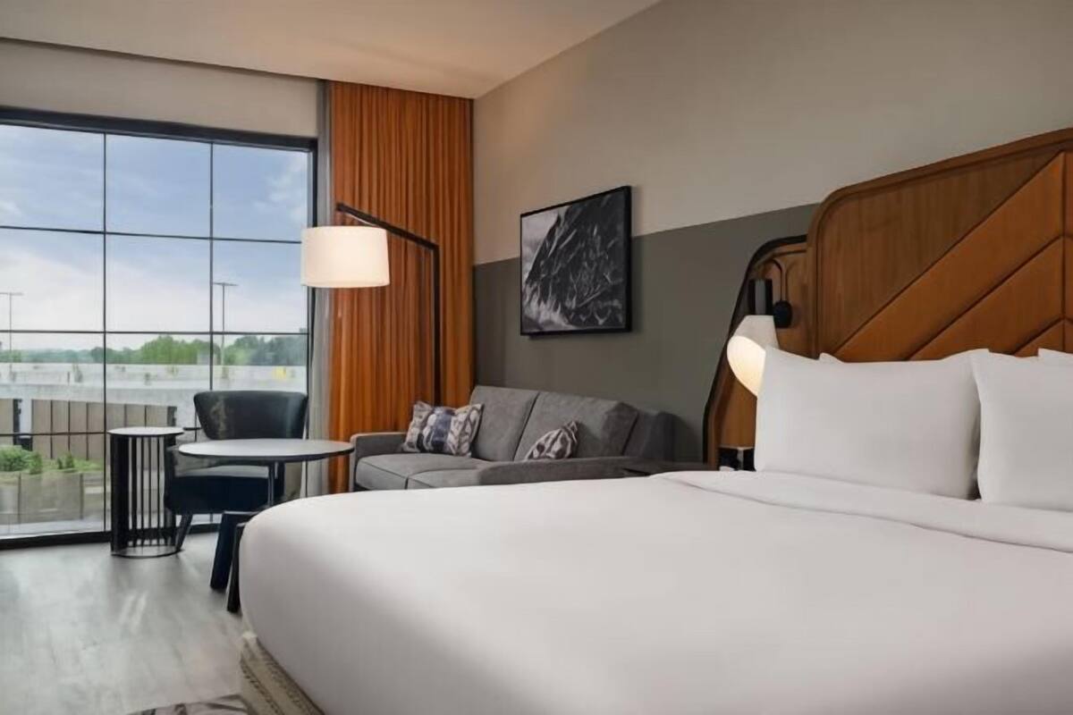 A hotel room with a large bed, a sofa, a small table, a chair, a large window, and a painting on the wall.
