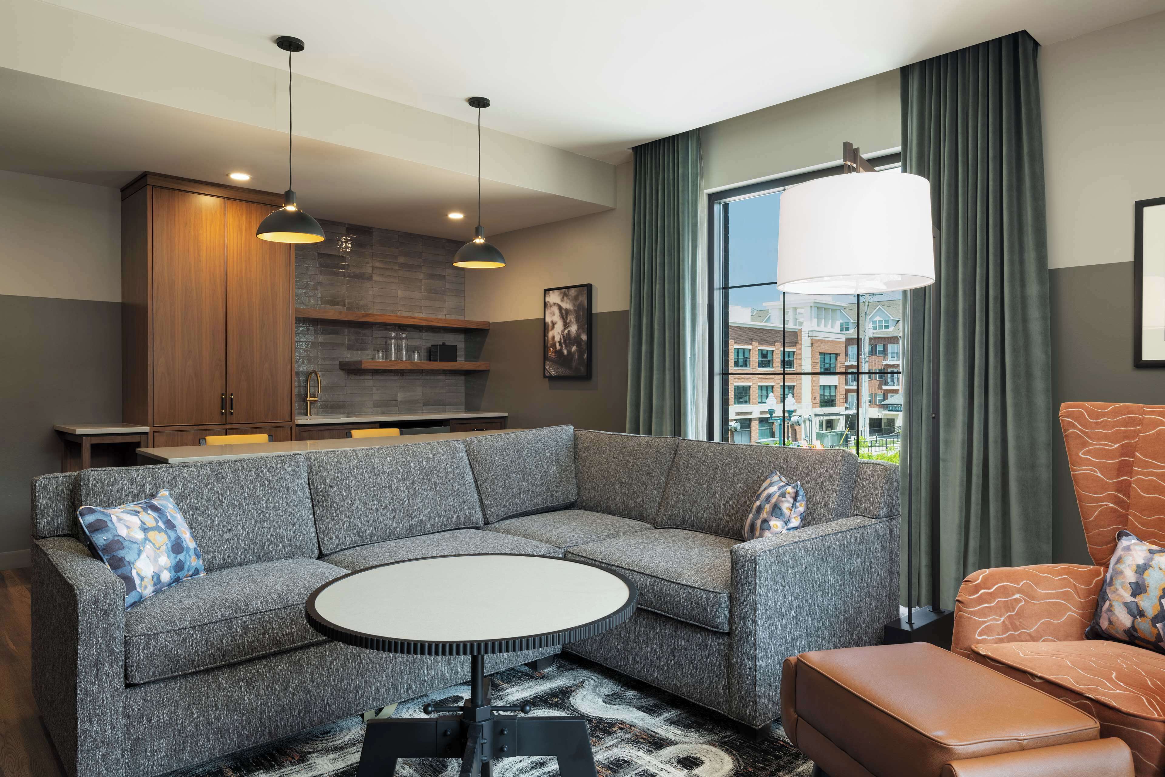 A modern living room with a gray sectional sofa, a round coffee table, a brown leather ottoman, and a kitchen area with wooden cabinets and a brick backsplash.