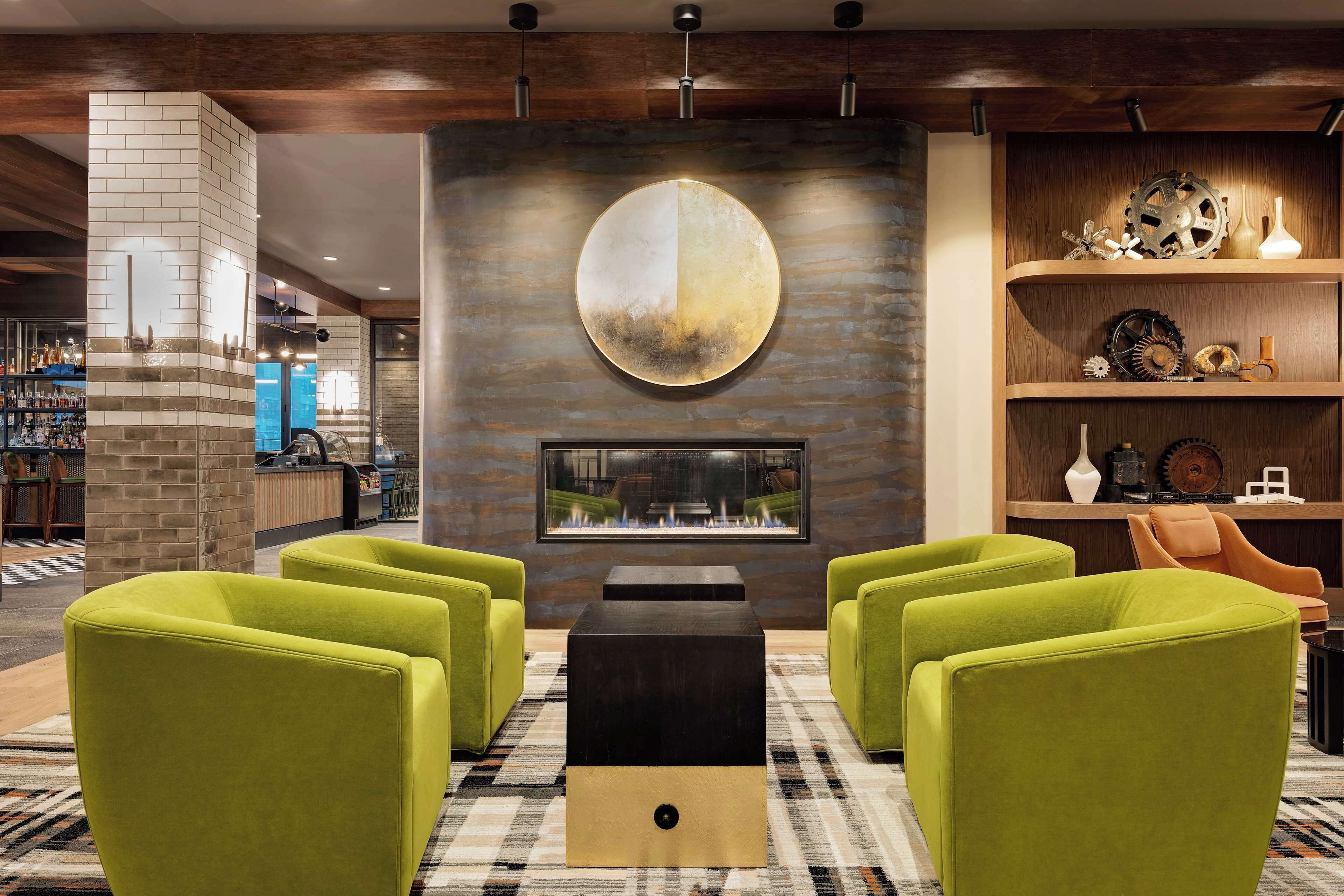 A modern lobby with a large circular wall art piece, a fireplace, and green armchairs.
