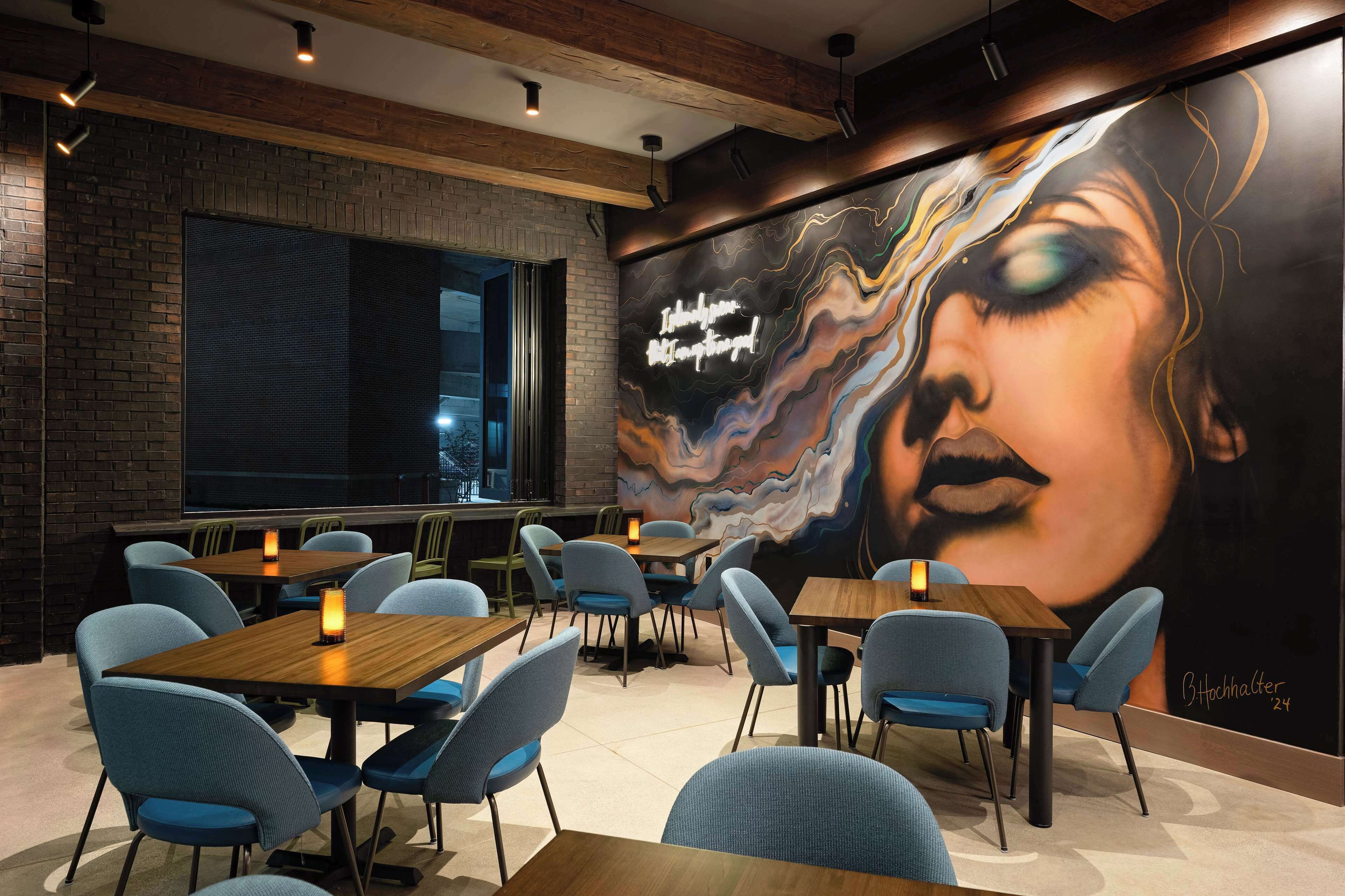 A restaurant interior with brick walls, blue chairs, wooden tables, and a large mural on the wall.