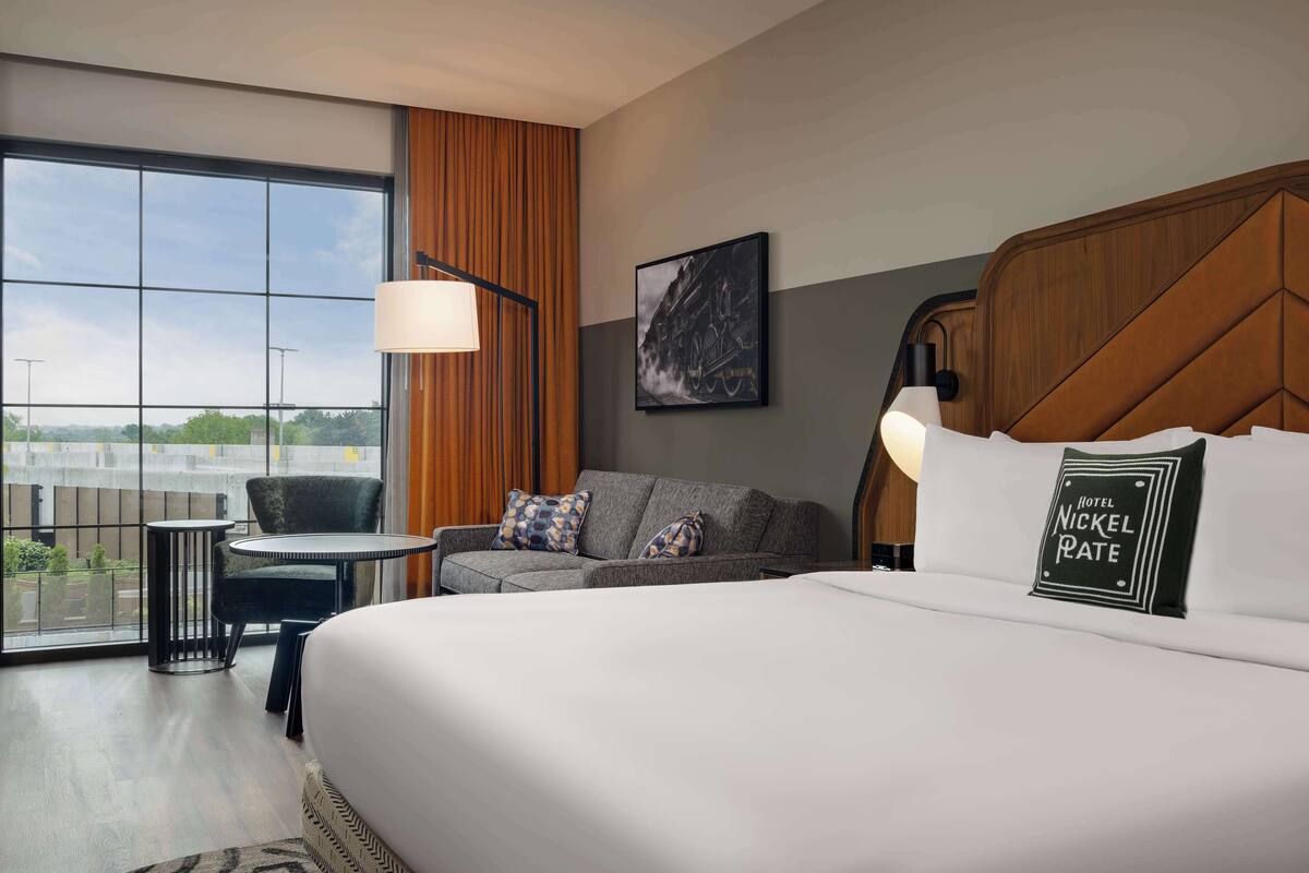 A hotel room with a large bed, a sofa, a small table, and a chair. There is a window with a view of the outside.