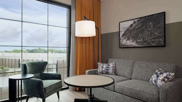 A modern living room with a grey sofa, a round table, and a green armchair. A large window offers a view of the cityscape.