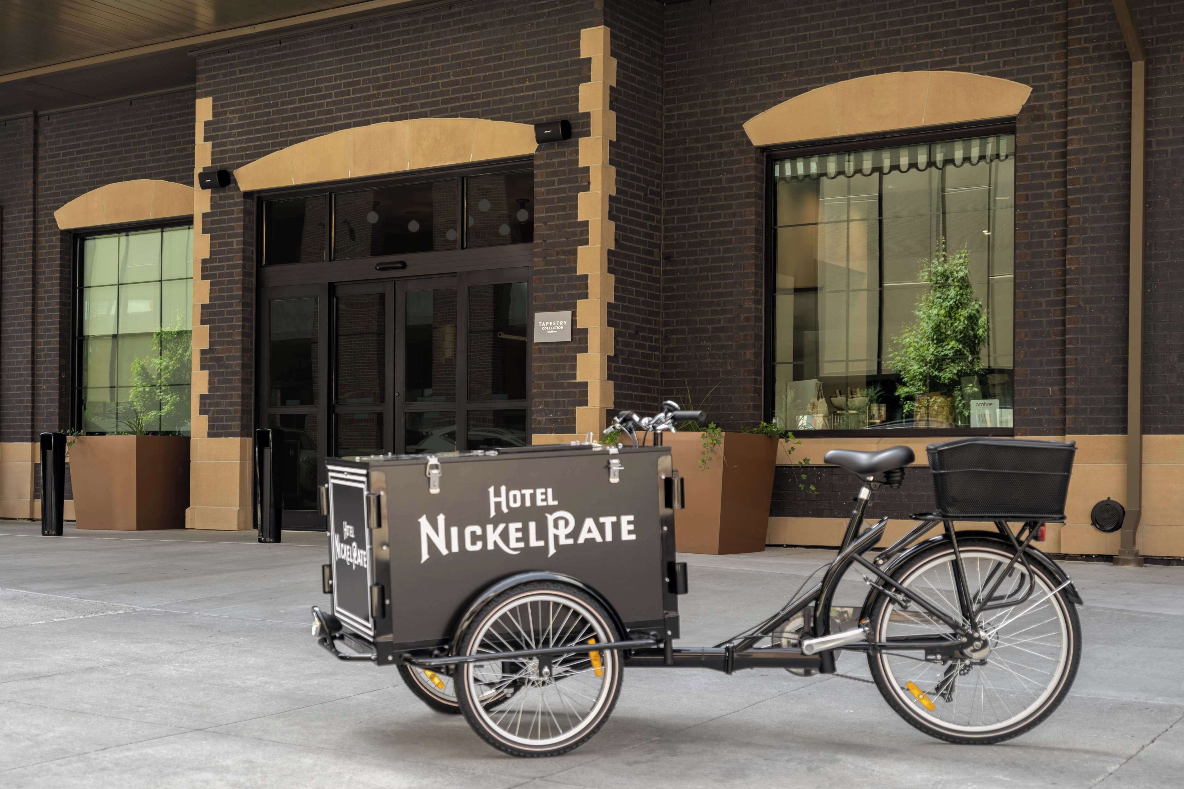 A bicycle with a trailer labeled "Hotel NickelPlate" parked in front of a building with large windows and a door.