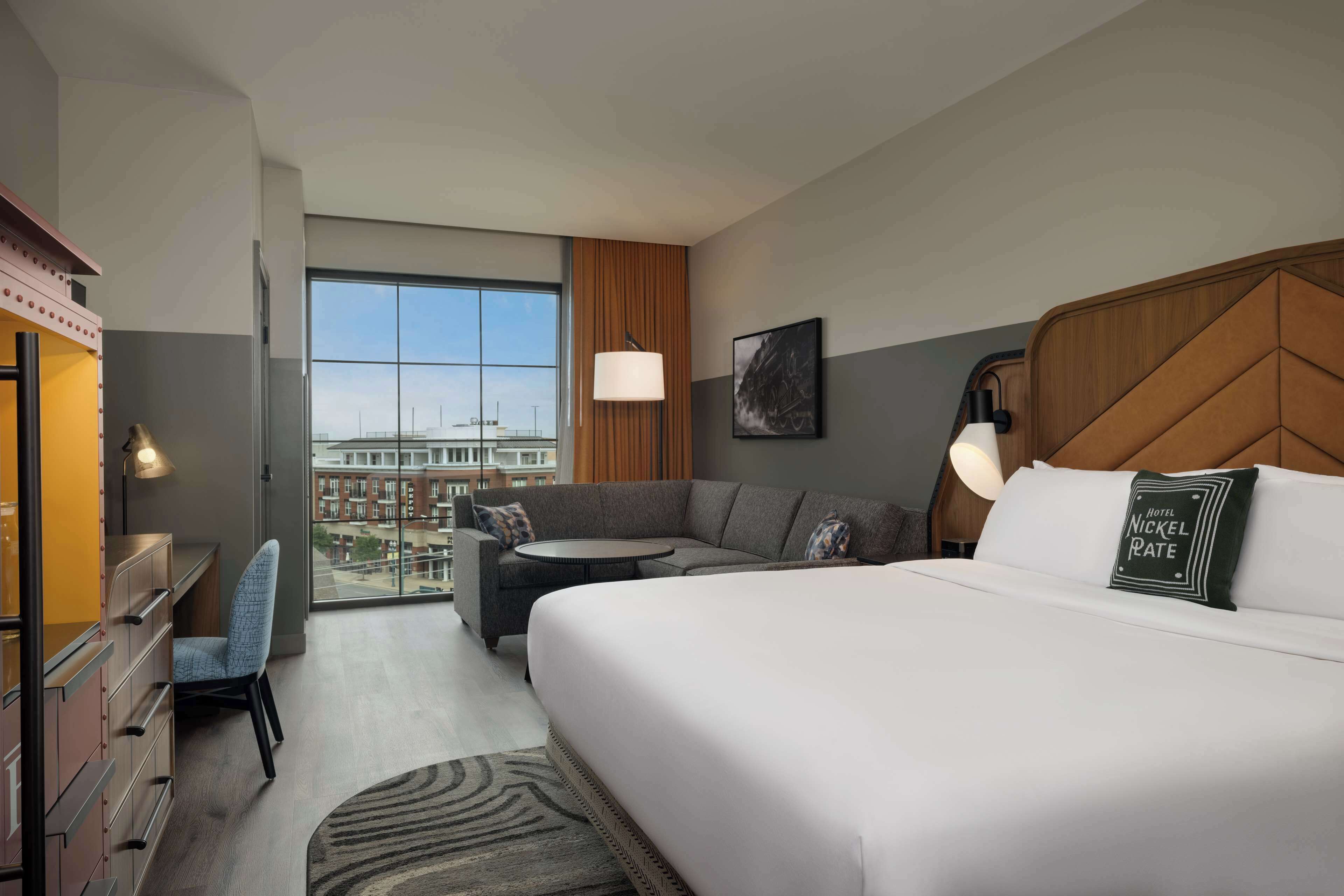 A hotel room with a large bed, a sofa, a desk, and a chair. There is a window with a view of buildings.