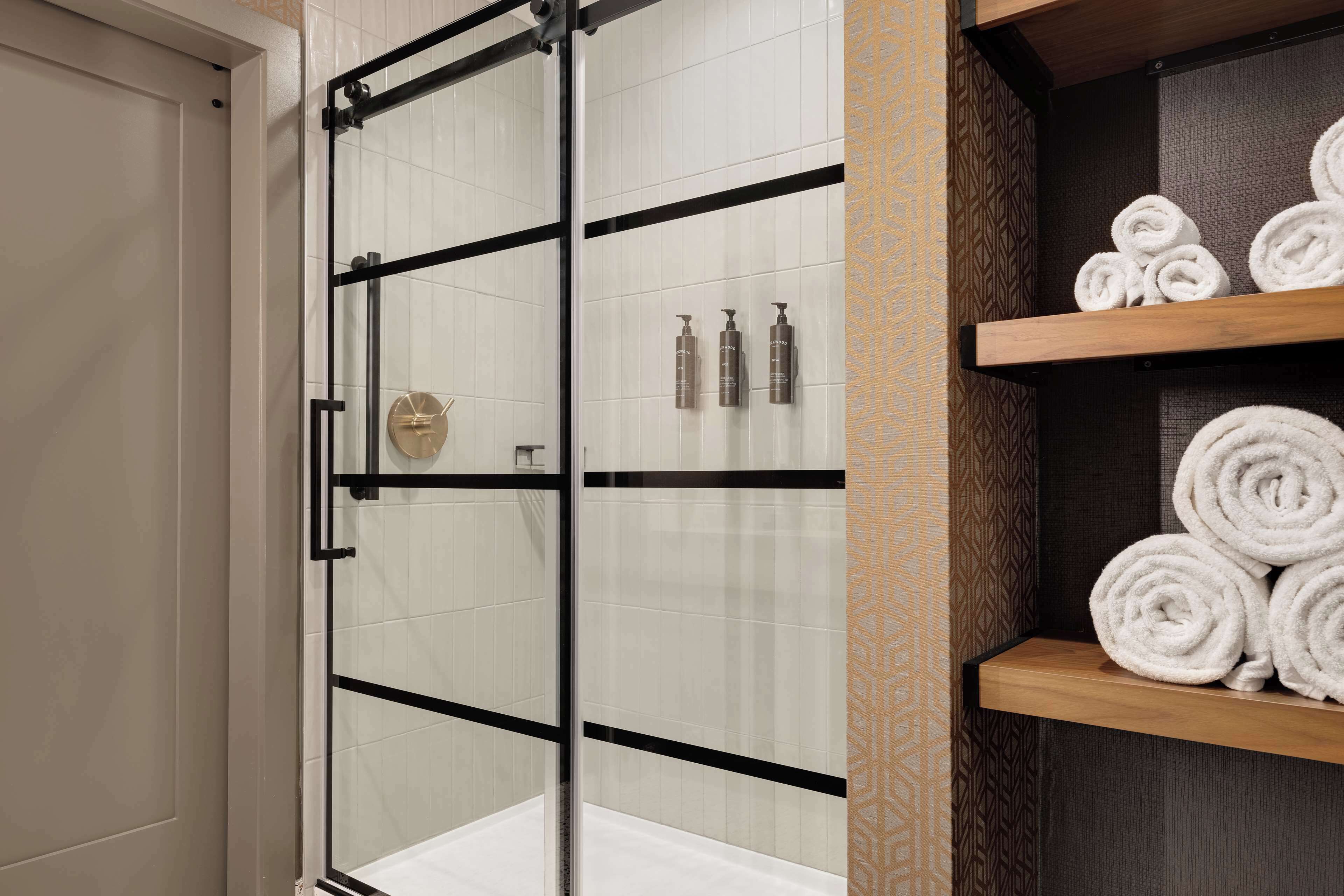A glass-enclosed shower with a black frame, white tiles, and a wooden shelf with rolled towels.