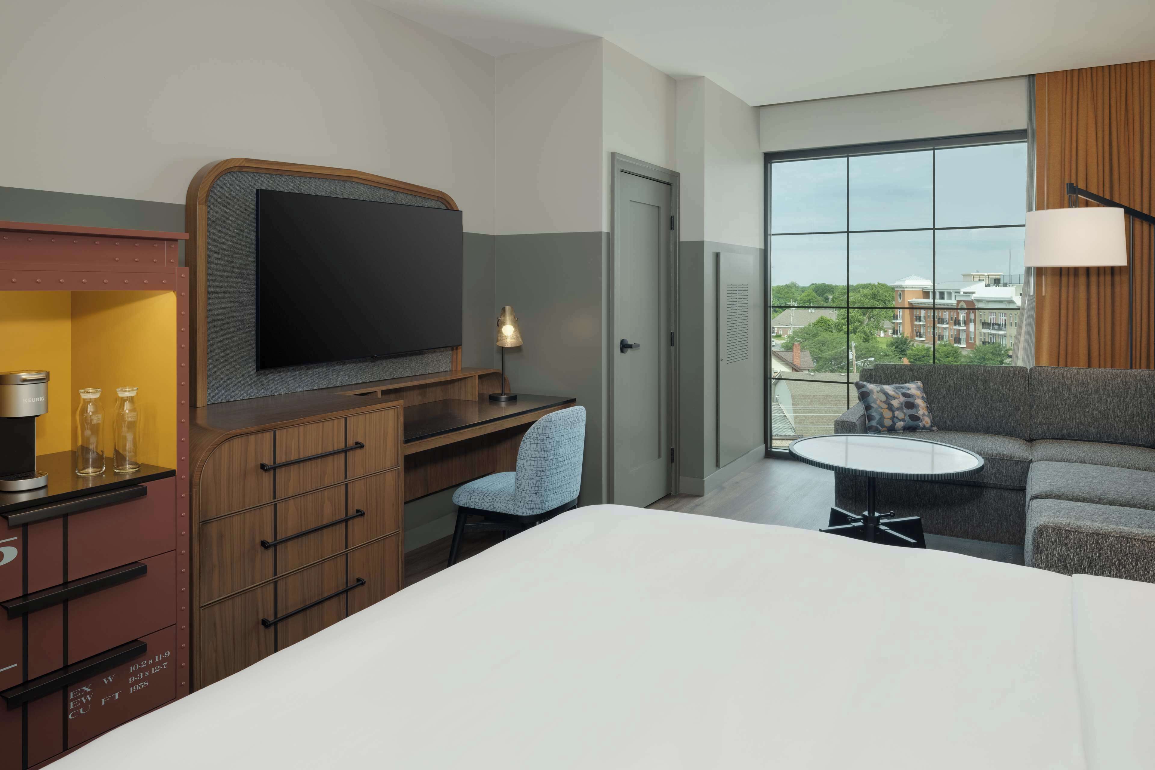Ein Hotelzimmer mit Bett, Schreibtisch mit Flachbildfernseher, Sessel, Sofa, kleinem Tisch und Fenster mit Blick auf die Stadt.