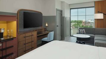 Ein Hotelzimmer mit Bett, Schreibtisch mit Flachbildfernseher, Sessel, Sofa, kleinem Tisch und Fenster mit Blick auf die Stadt.