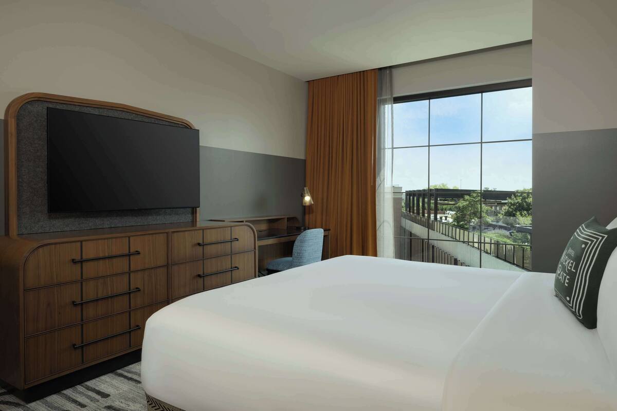 A hotel room with a large bed, a TV mounted on a wooden dresser, a desk with a chair, and a large window offering a view of the city.