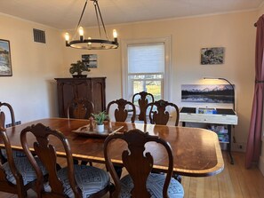 A dining room with a wooden table, chairs, a chandelier, a window, and a piano. - Wagon Wheel Farm House at Explore Acadia Cottages (Bar Harbor)