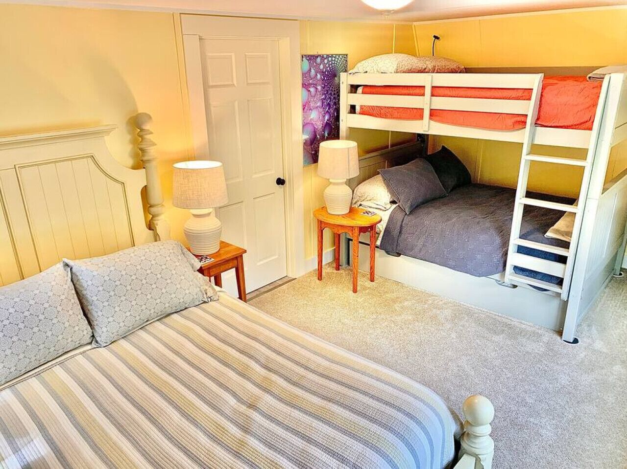 Une chambre avec un lit superposé, un lit simple, une lampe, une petite table et une porte.