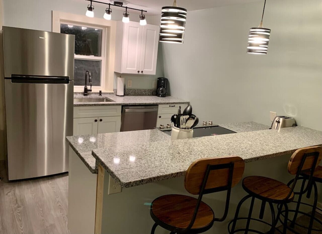 A modern kitchen with a stainless steel refrigerator, granite countertop, bar stools, and pendant lighting.