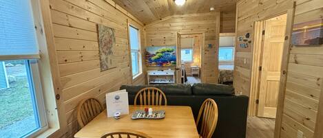 A wooden cabin interior with a dining table, chairs, a sofa, and a painting on the wall.