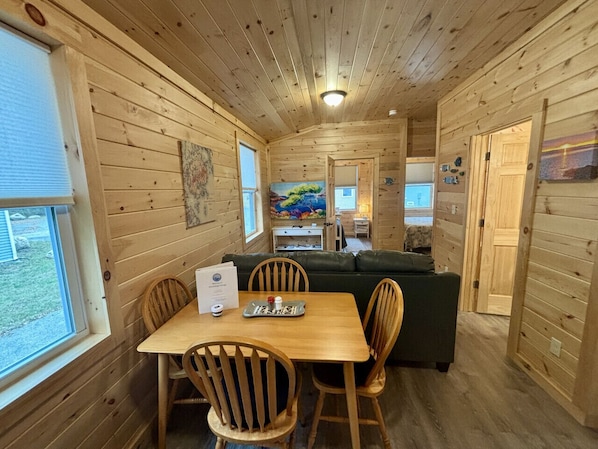 A wooden cabin interior with a dining table, chairs, a sofa, and a painting on the wall.