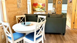 A wooden cabin interior with a dining table, chairs, a sofa, and a painting on the wall. - Hadley Point Cottage #1 at Explore Acadia Cottages (Bar Harbor)