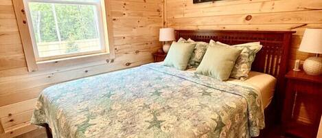 Un dormitorio de cabaña de madera con una cama, dos mesitas de noche, un ventanal y un cuadro en la pared.