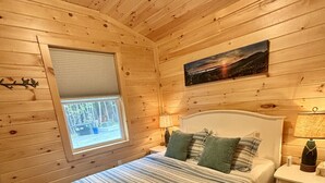 A cozy wooden room with a bed, a window, a nightstand, and a painting on the wall.