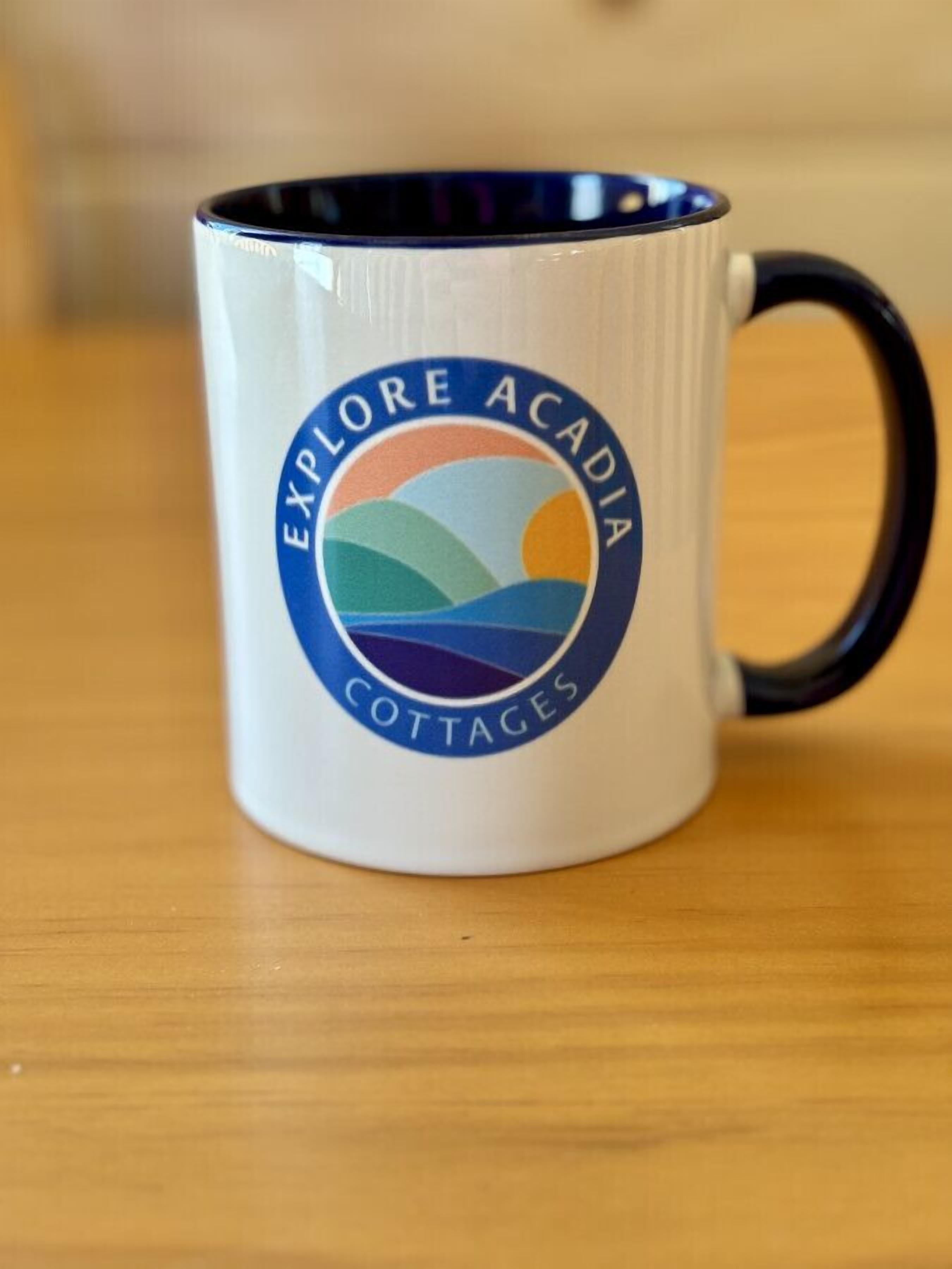 A white mug with a logo that reads "EXPLORE ACADIA COTTAGES".
