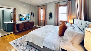Une chambre à coucher comprenant un lit, un ventilateur de plafond, une télévision, une commode et une fenêtre avec des rideaux.