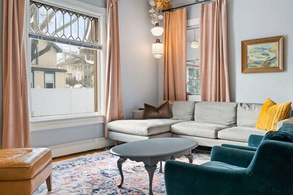 A living room with a sectional sofa, a round coffee table, a small ottoman, and a window with curtains.
