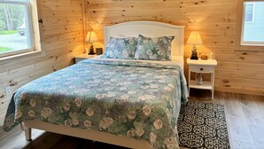 A wooden cabin bedroom with a bed, two bedside tables, a nightstand, a window, and a wall-mounted picture. - Sea Star Cottage #6 at Explore Acadia Cottages (Bar Harbor)