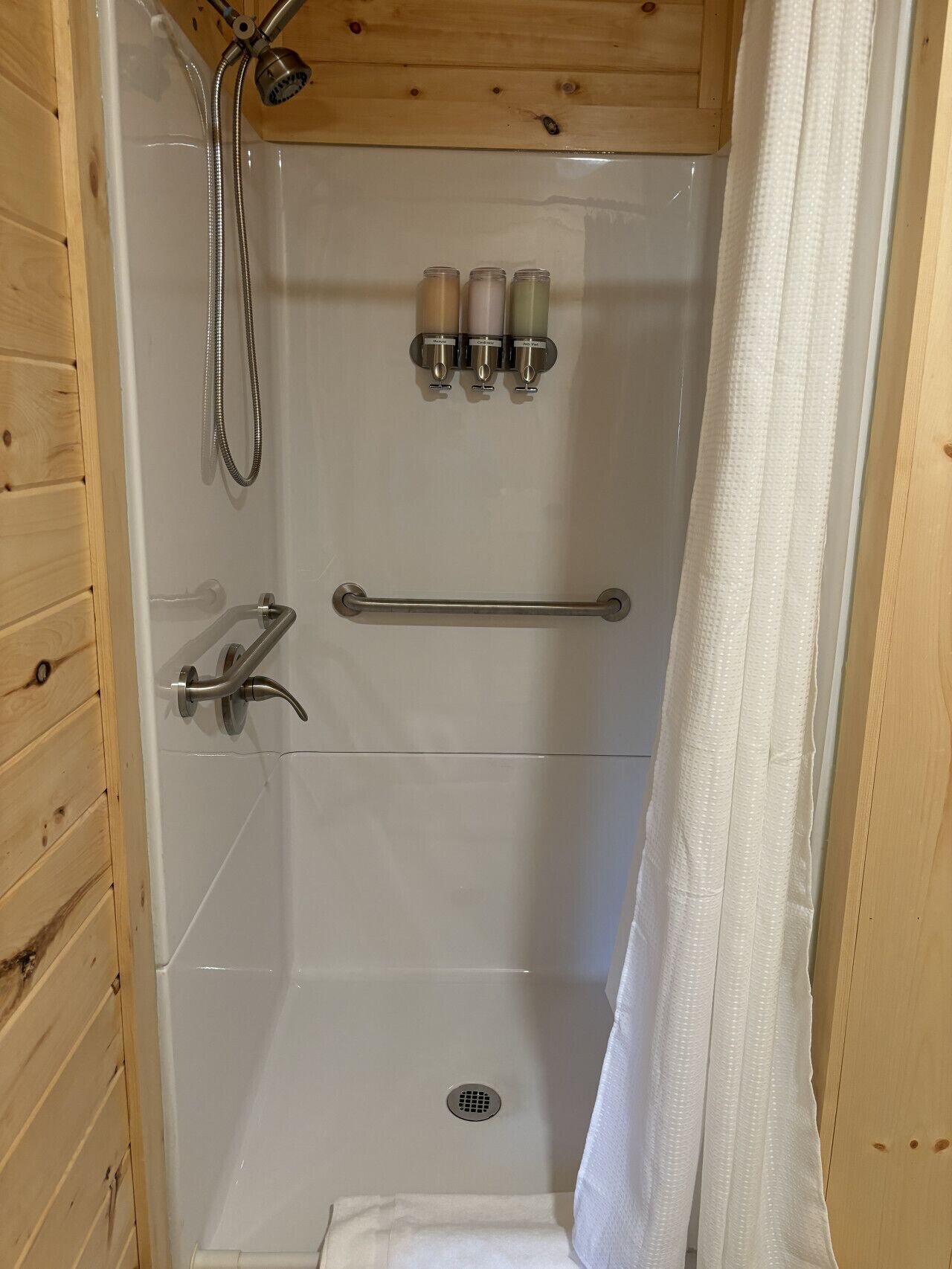 A shower stall with a wooden ceiling, a handrail, a showerhead, and three bottles of shampoo.