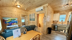 A wooden cabin interior with a dining table, chairs, a sofa, a kitchen area, and a painting on the wall. - Sea Star Cottage #6 at Explore Acadia Cottages (Bar Harbor)