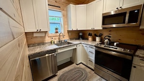 A kitchen with wooden walls, white cabinets, stainless steel appliances, and a window with a view. - Sea Star Cottage #6 at Explore Acadia Cottages (Bar Harbor)