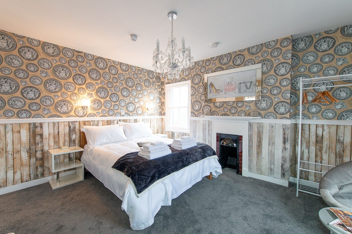 A bedroom with a bed, a chandelier, a fireplace, and patterned wallpaper.