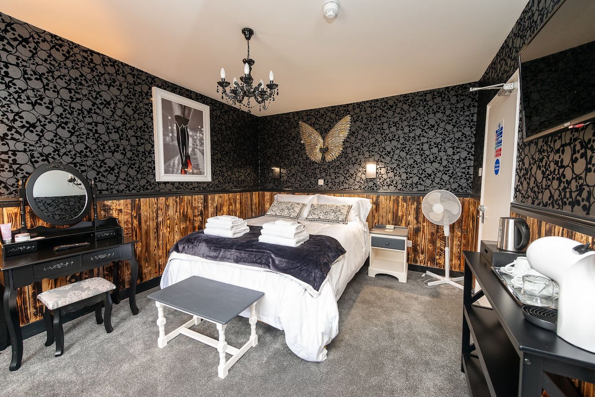 A bedroom with a bed, bedside tables, a mirror, a bench, a fan, and a wall-mounted decorative piece.