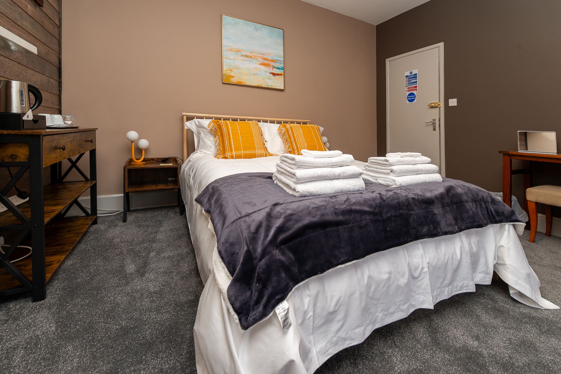 A neatly arranged hotel room with a bed, a nightstand, a desk, and a door with a sign.