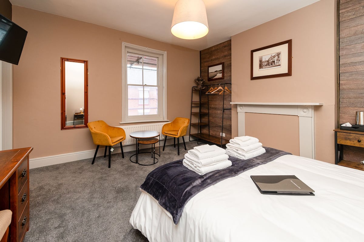 A hotel room with a bed, a desk, two chairs, a coat rack, and a fireplace.