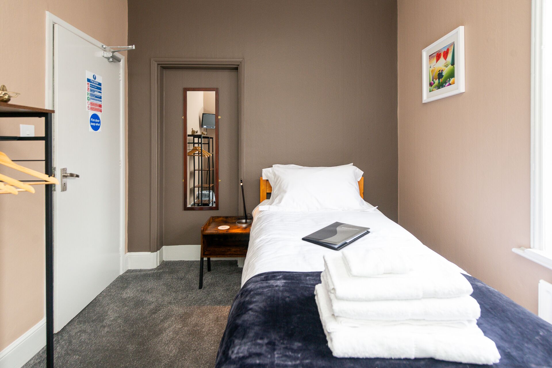 Une petite chambre à coucher, bien rangée, avec un lit simple, une table de chevet, une chaise, un miroir et une porte.
