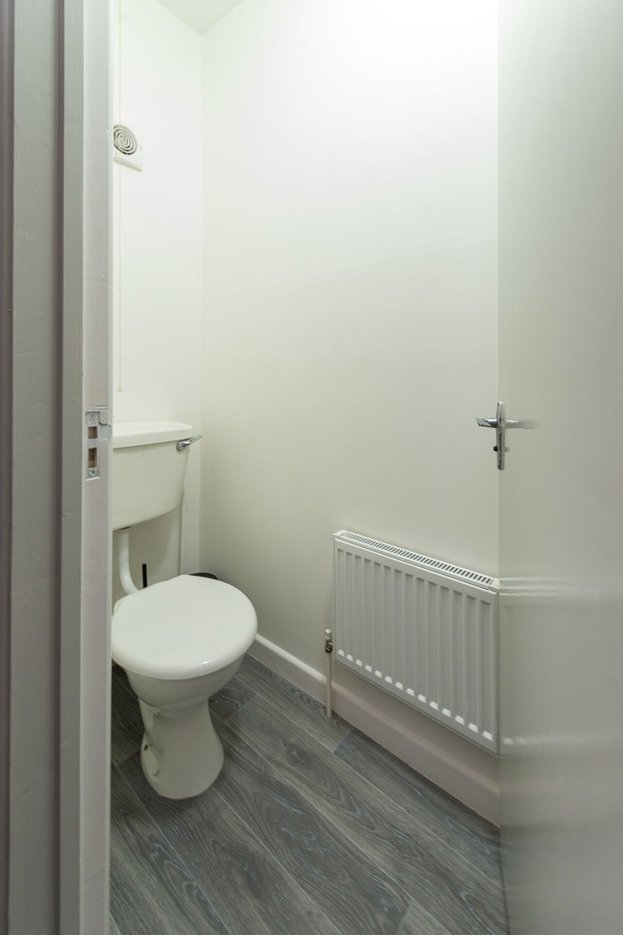 A small, white bathroom with a toilet, a white radiator, and a grey wooden floor.
