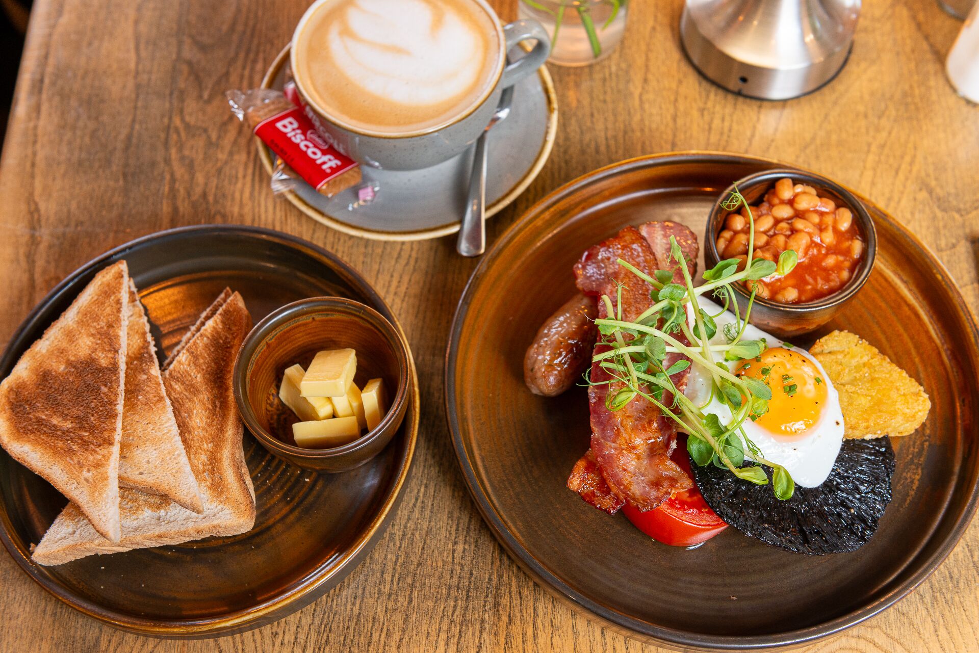 A breakfast setting with toast, butter, sausages, beans, a fried egg, and a cup of coffee on a wooden table.