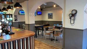 A bar area with wooden counter and hanging lights, tables and chairs, and a TV displaying sports. - The Bosun's Chair (Lymington)