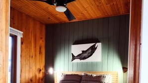 Ein Schlafzimmer mit einem Bett, Holzboden, einem Deckenventilator, einem Wandbild mit Fischen und einem Fenster.