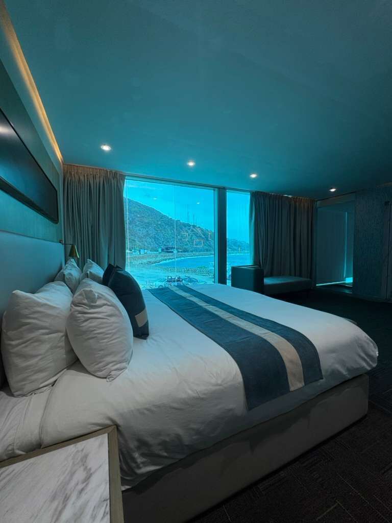 A large bed with multiple pillows, a bedside table, and a view of a mountainous landscape through large windows.