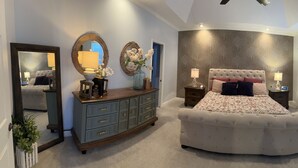 Ein Schlafzimmer mit einem großen Bett, einem Schminktisch, einem Fernseher und einem durchgehenden Türrahmen sichtbaren Badezimmer.