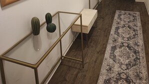 Un pasillo con suelo de madera, un tapete estampado, una mesa de vidrio sobre patas de metal, un banco y dos cuadros en la pared.