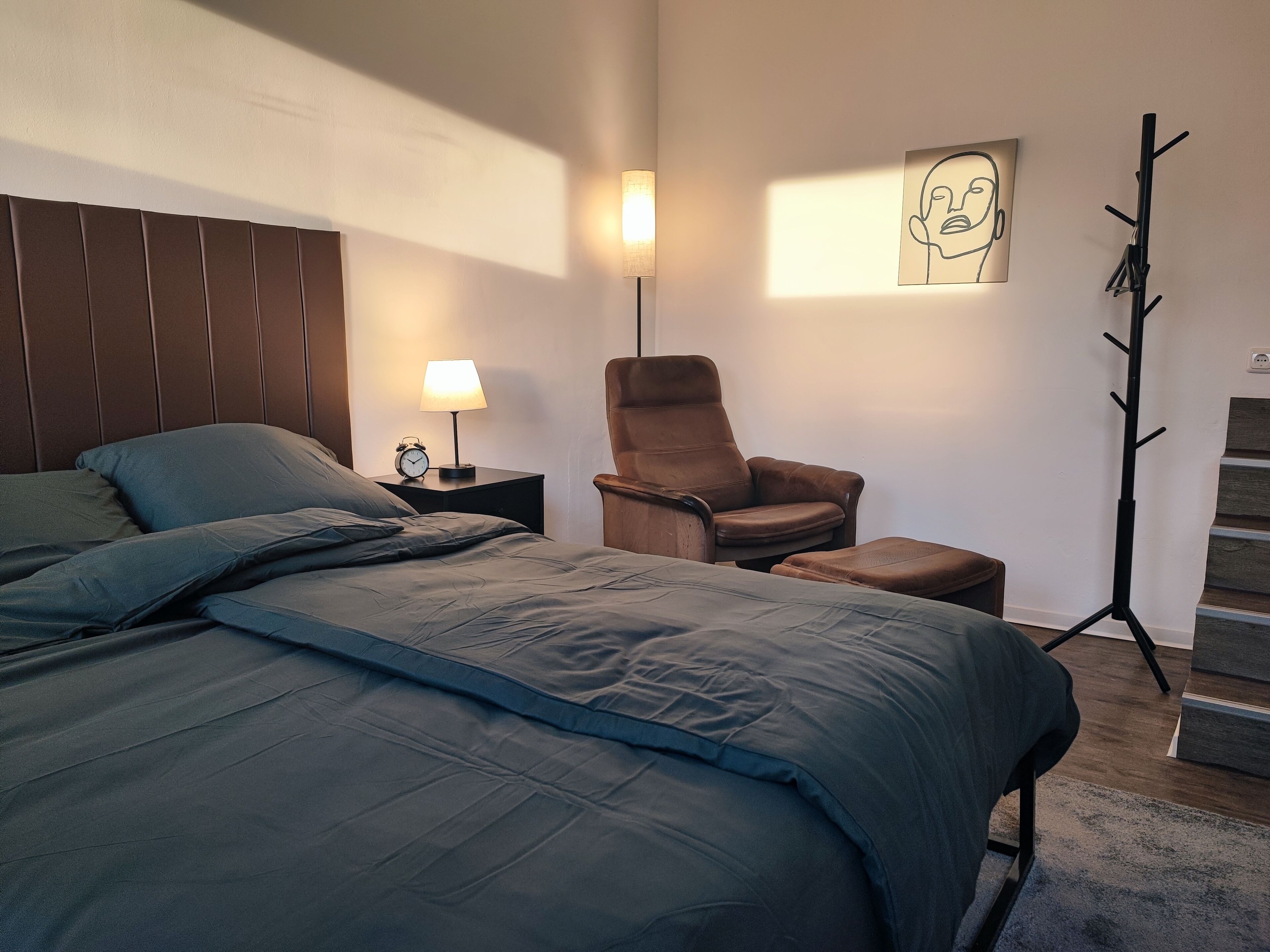 A bedroom with a bed, a chair, a nightstand with a lamp, a coat rack, and a painting on the wall.