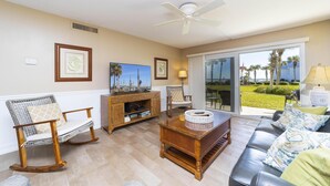Sala de estar con una mecedora de madera, un sofá, una mesa de centro y un televisor. Se ve una ventana con vistas a palmeras y un jardín.