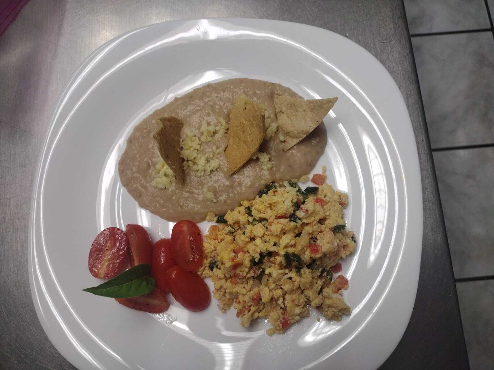 Un plato con una porción generosa de hummus, una rebanada de pan, una guarnición de huevos revueltos y tomates en rodajas.