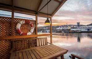 Un banco de madera y un círculo salvavidas colgados en un muro de celosía, con vistas a un río con barcos y edificios al fondo.