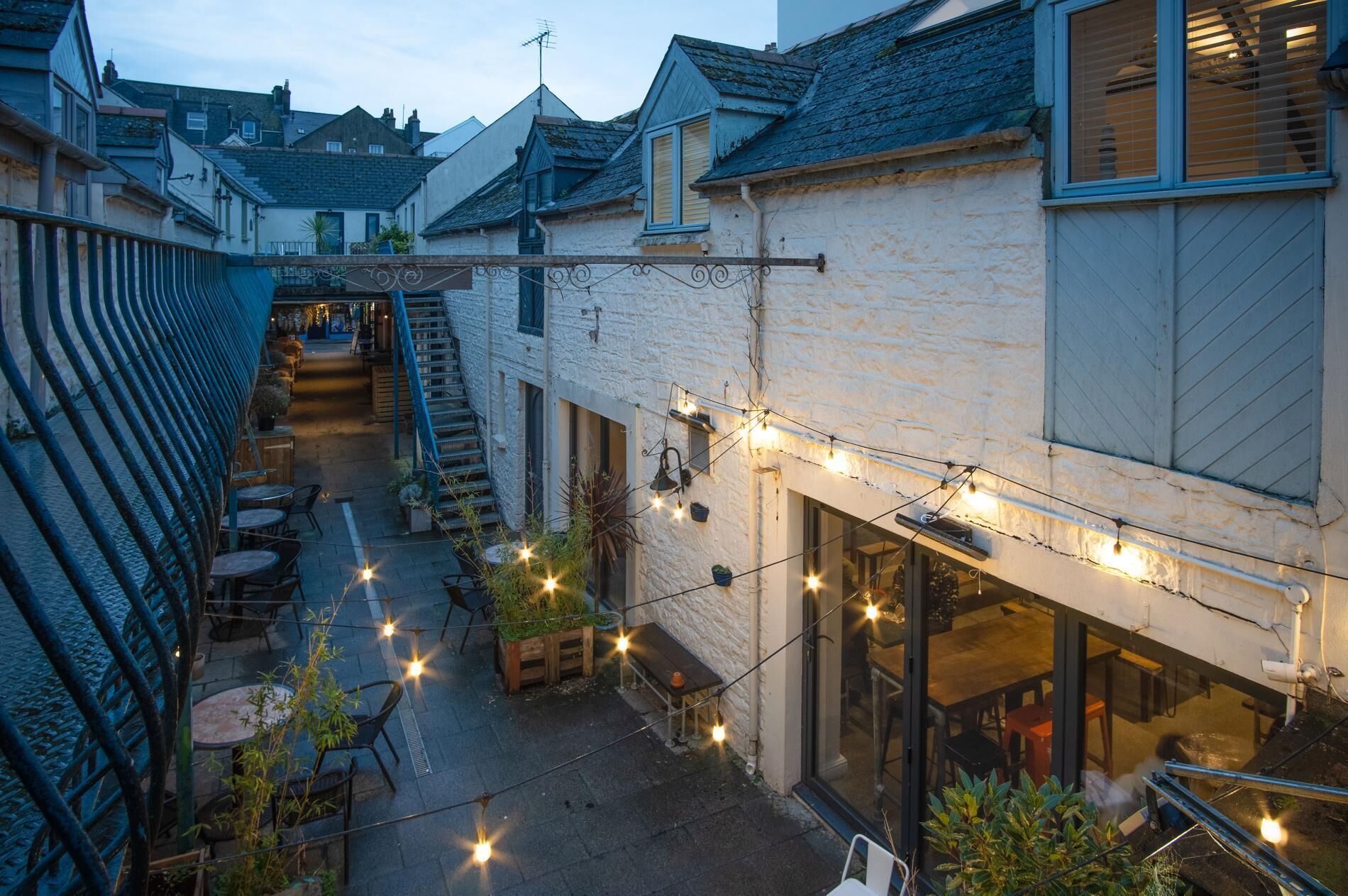 Une ruelle étroite avec un restaurant sur la droite, doté d’une terrasse et illuminée de guirlandes lumineuses.
