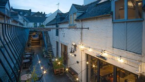 Un estrecho callejón con un restaurante a la derecha, que ofrece mesas al aire libre y luces colgantes.