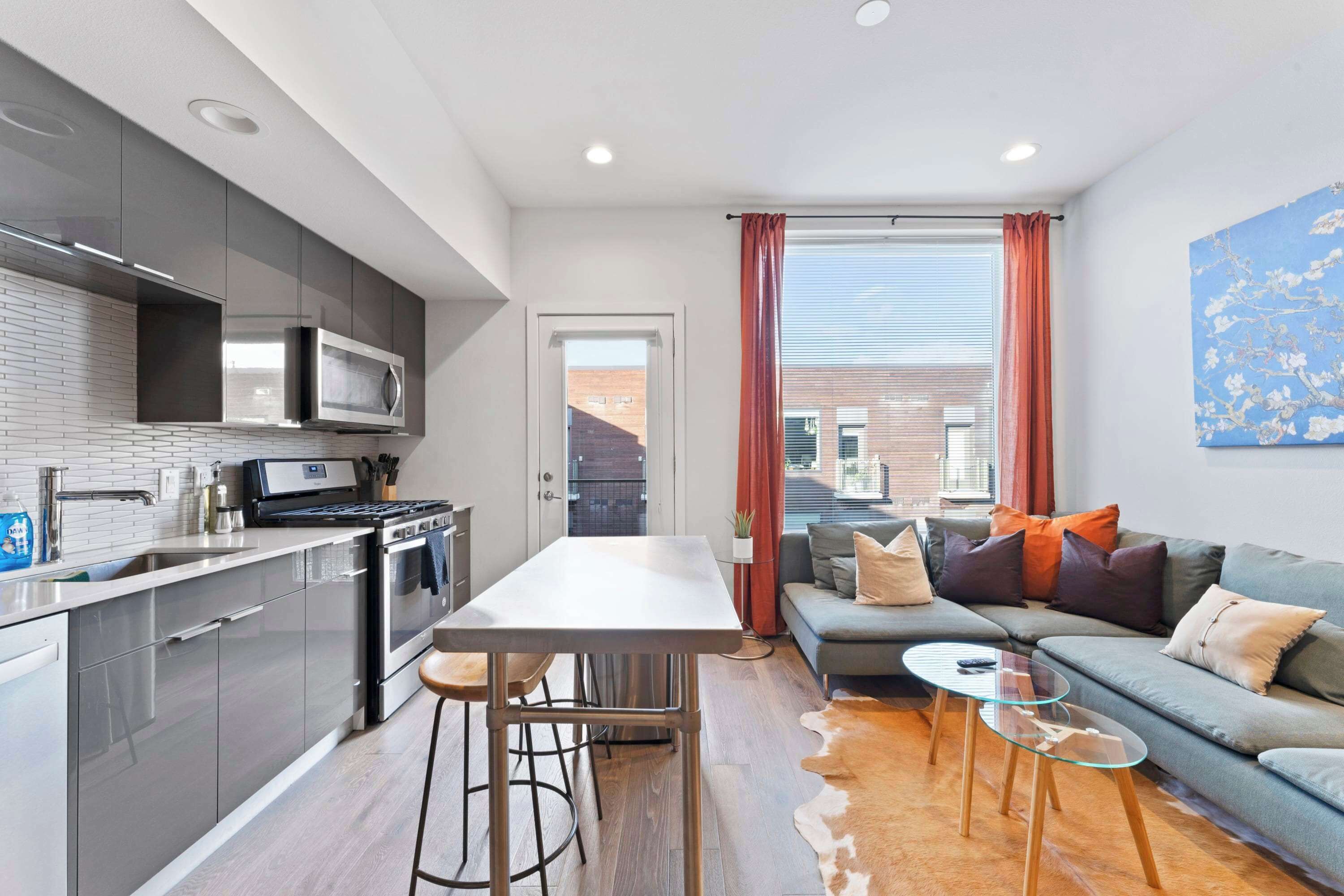 A modern kitchen with stainless steel appliances, a dining area with stools, and a living room with a sofa and a glass coffee table.