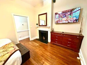 A bedroom with a bed, a dresser, a mirror, a fireplace, and a television mounted on the wall.
