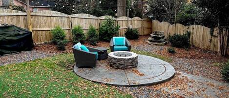 A circular outdoor seating area with a stone fire pit, surrounded by a wooden fence, and a grassy lawn with scattered leaves.
