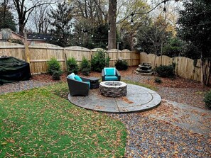 A circular outdoor seating area with a stone fire pit, surrounded by a wooden fence, and a grassy lawn with scattered leaves.