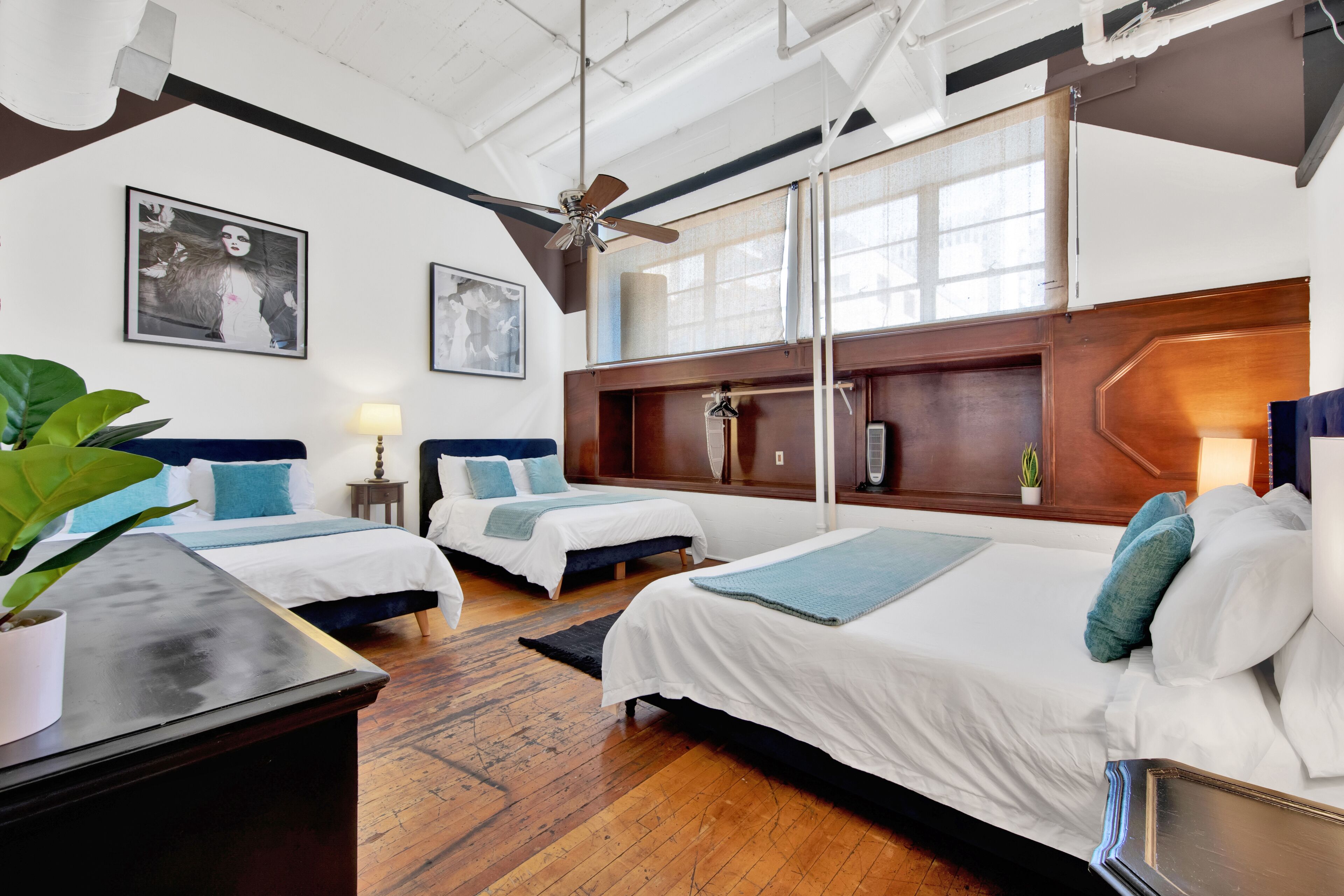 A hotel room with two beds, a ceiling fan, large windows, and artwork on the walls.